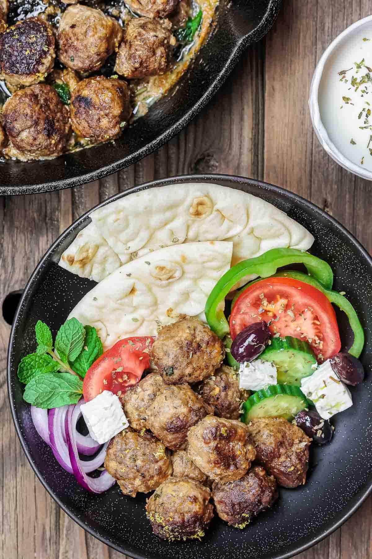 Greek meatballs (keftedes) in a bowl with mint, red onions, pita bread, olives, feta cheese, cucumber, tomato, and green bell peppers.