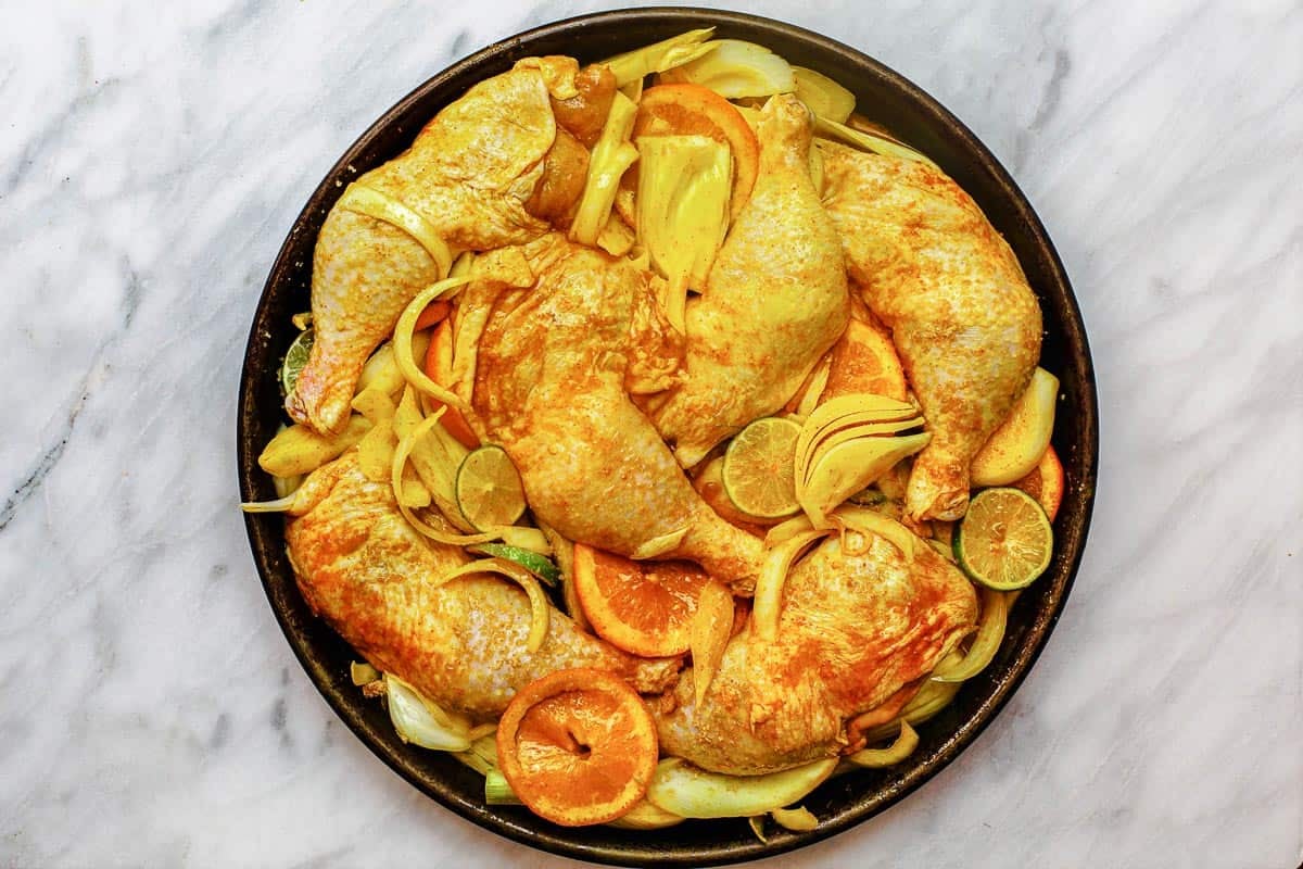chicken leg quarters in a round baking dish with onions, fresh fennel, lime slices, and orange slices.