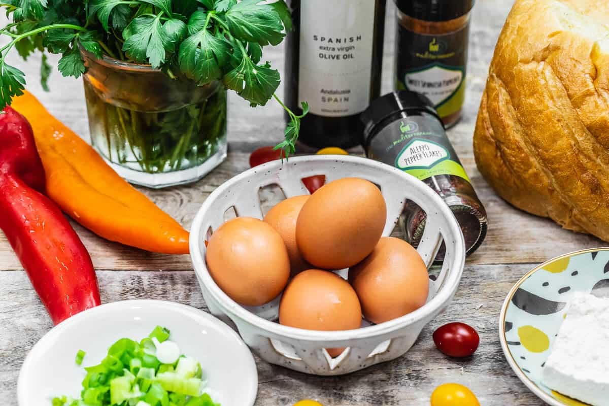 ingredients for egg toast including parsley, olive oil, spices, bread, feta cheese, grape tomatoes, eggs, green onions, and peppers.