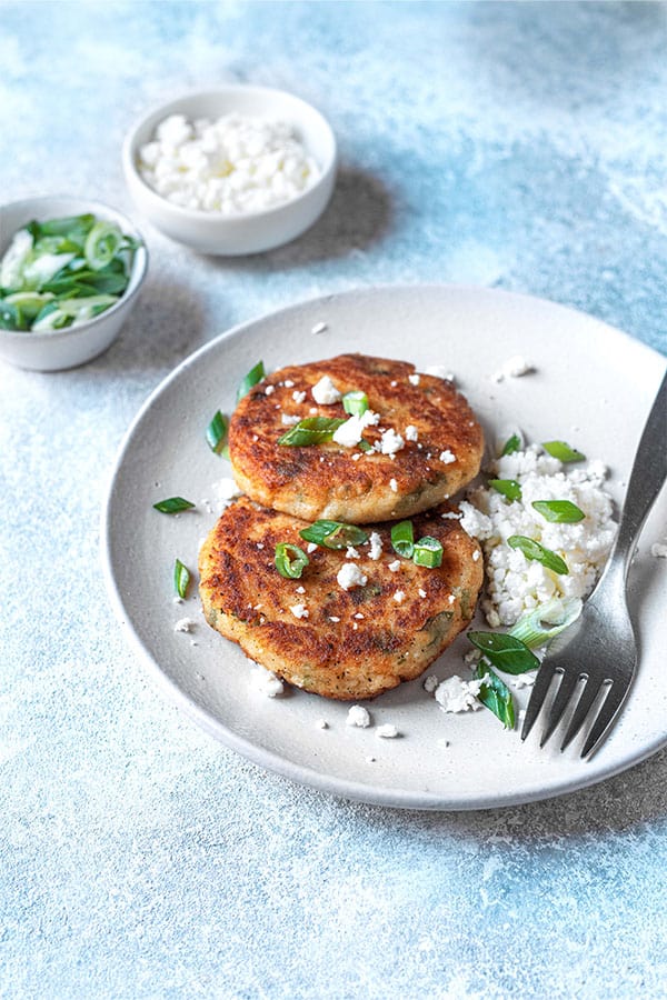 two mashed potato pancakes on a plate with a fork and scallions and feta cheese in the background.
