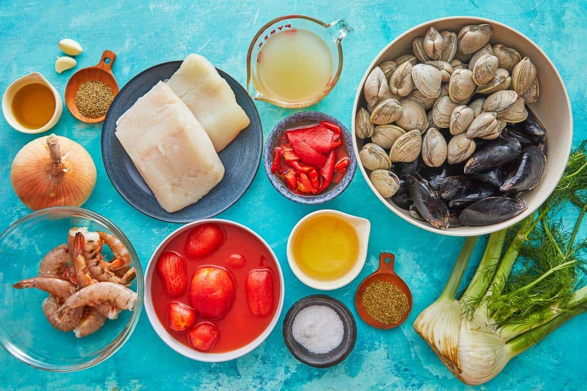 ingredients for cioppino including clams, mussels, fennel, olive oil, onion, kosher salt, garlic, roasted red peppers, whole tomatoes, thyme, oregano, white wine, seafood stock, white fish, and shrimp.