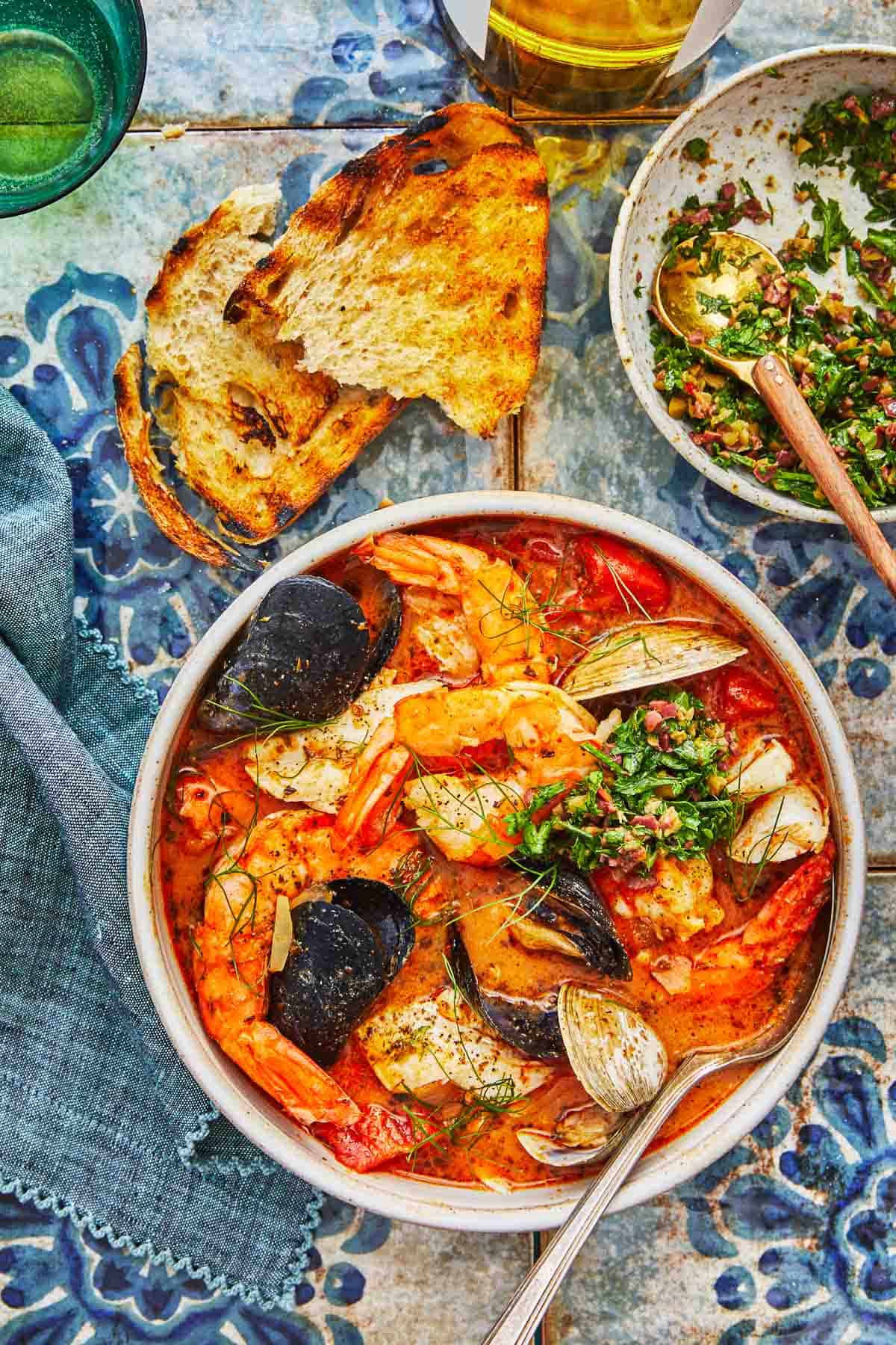 a bowl of cioppino with a spoon, two pieces of toasted and a bowl of Parsley-Olive Gremolata.