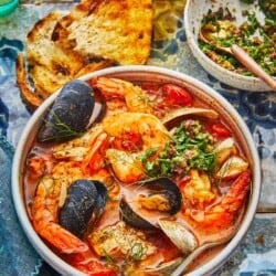 a bowl of cioppino with a spoon, two pieces of toasted and a bowl of Parsley-Olive Gremolata.