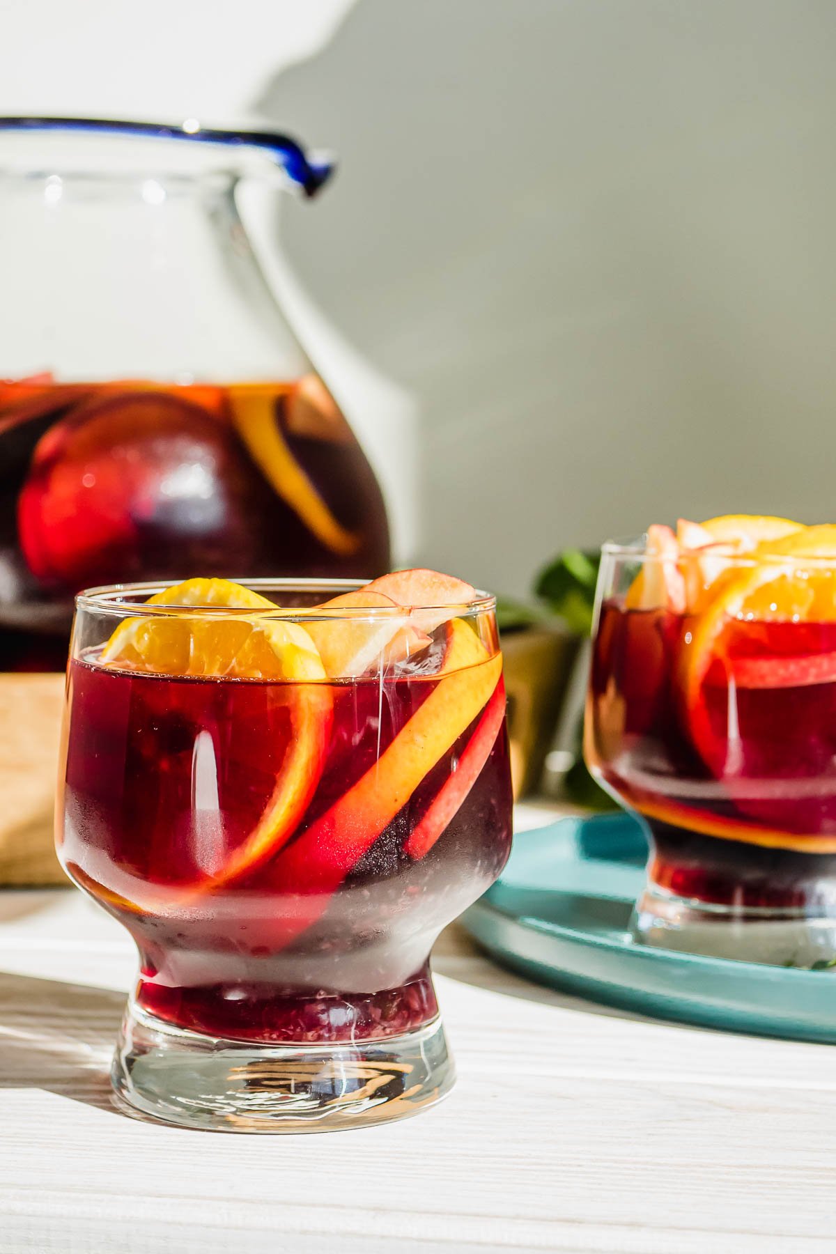 two glasses of red sangria and sliced oranges with a pitcher of sangria in the background.
