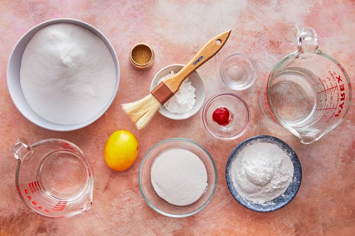 ingredients for turkish delight (lokum) including powdered sugar, cornstarch, superfine sugar, cold water, lemon, powdered gelatin, rose water, red food coloring and gold edible glitter.