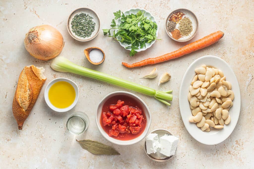 Ingredients for gigantes plaki (Greek baked beans) including olive oil, carrot, celery, garlic, kosher salt, tomatoes, parsley, thyme, spices, bay leaf, butter beans, feta cheese and crusty bread.