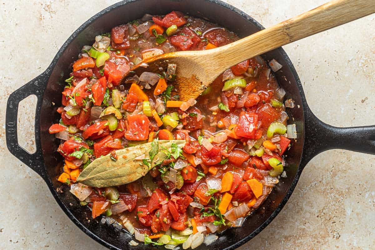 Greek baked white beans (gigantes plaki) topped with a bay leaf being cooked in a cast iron skillet with a wooden spoon.