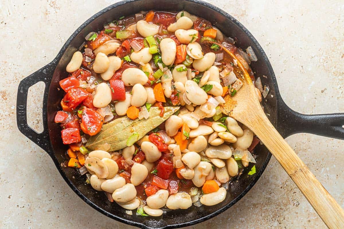 Greek baked white beans (gigantes plaki) topped with a bay leaf being cooked in a cast iron skillet with a wooden spoon.
