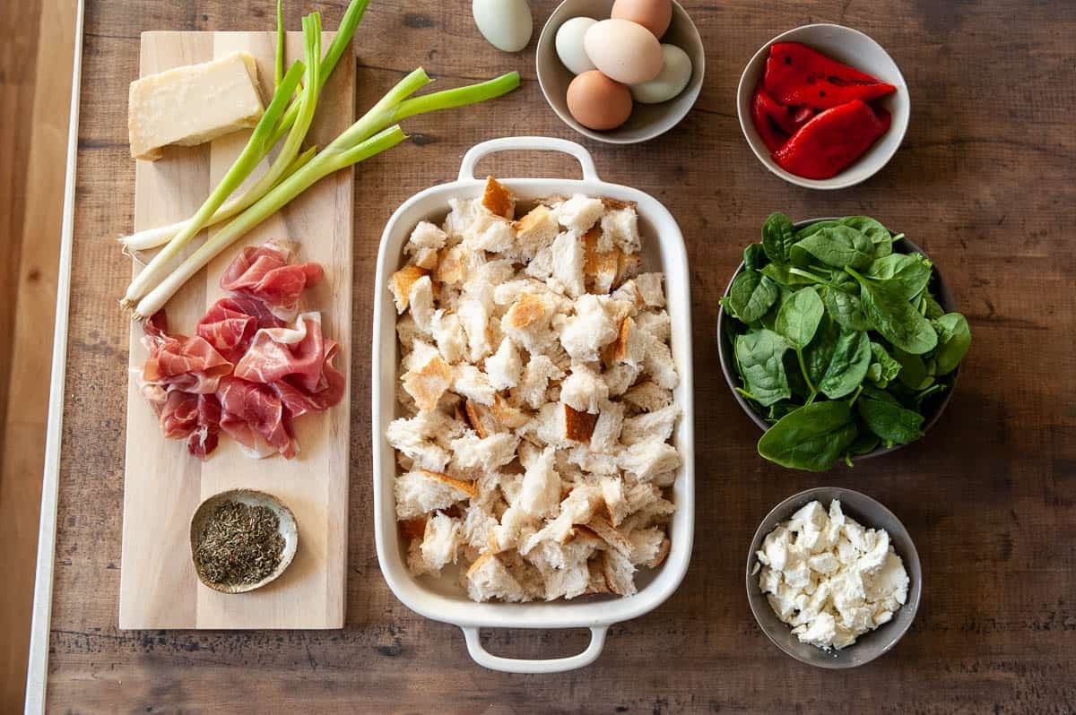 ingredients for breakfast strata including cubed bread, cheese, spinach, roasted red peppers, prosciutto, scallions, eggs, and italian seasoning.