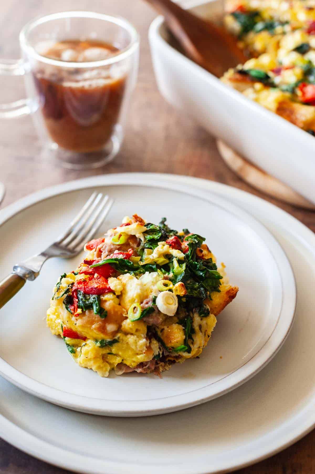 a slice of strata on a plate with a fork, and a mug of coffee in the background.