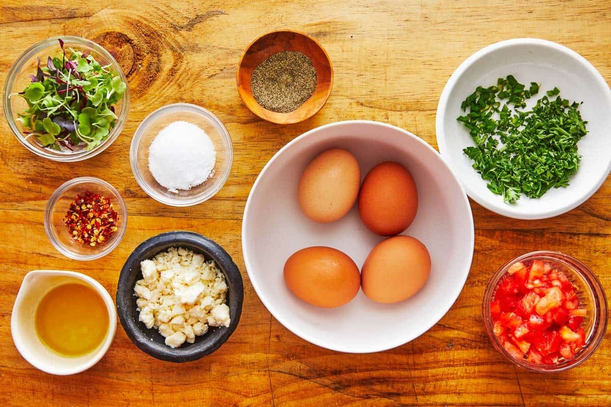 ingredients for baked eggs including olive oil, 4 eggs, salt, pepper, red pepper flakes, feta cheese, chives, parsley, microgreens and diced tomatoes.