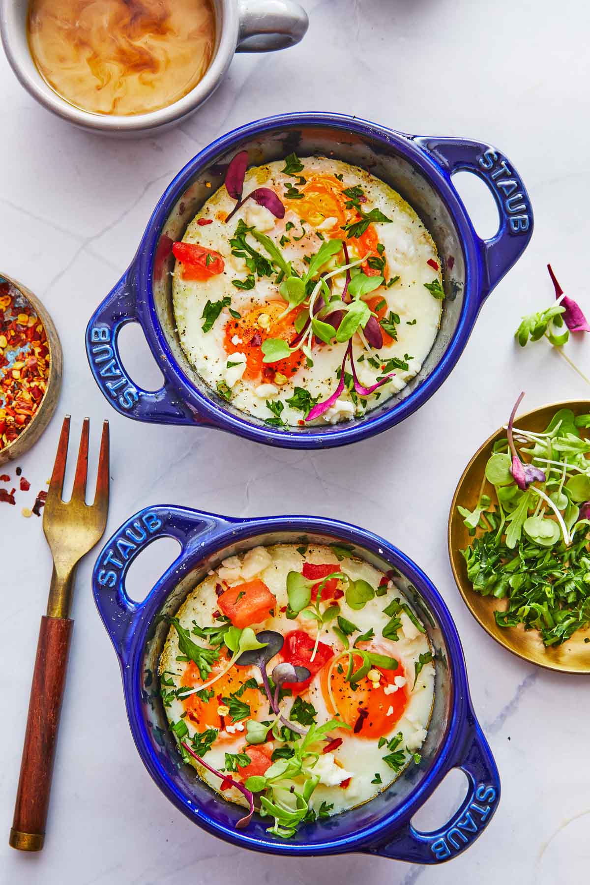 two small dishes with cooked eggs next to a fork, a cup of coffee, and a bowl of microgreens..