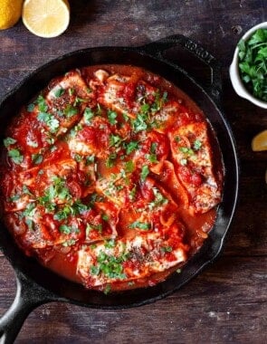 Greek fish with tomatoes and onions in a cast iron pan. parsley and lemon wedges to the side for serving