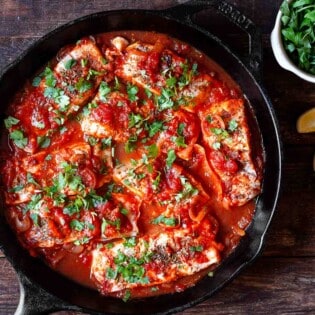Greek fish with tomatoes and onions in a cast iron pan. parsley and lemon wedges to the side for serving