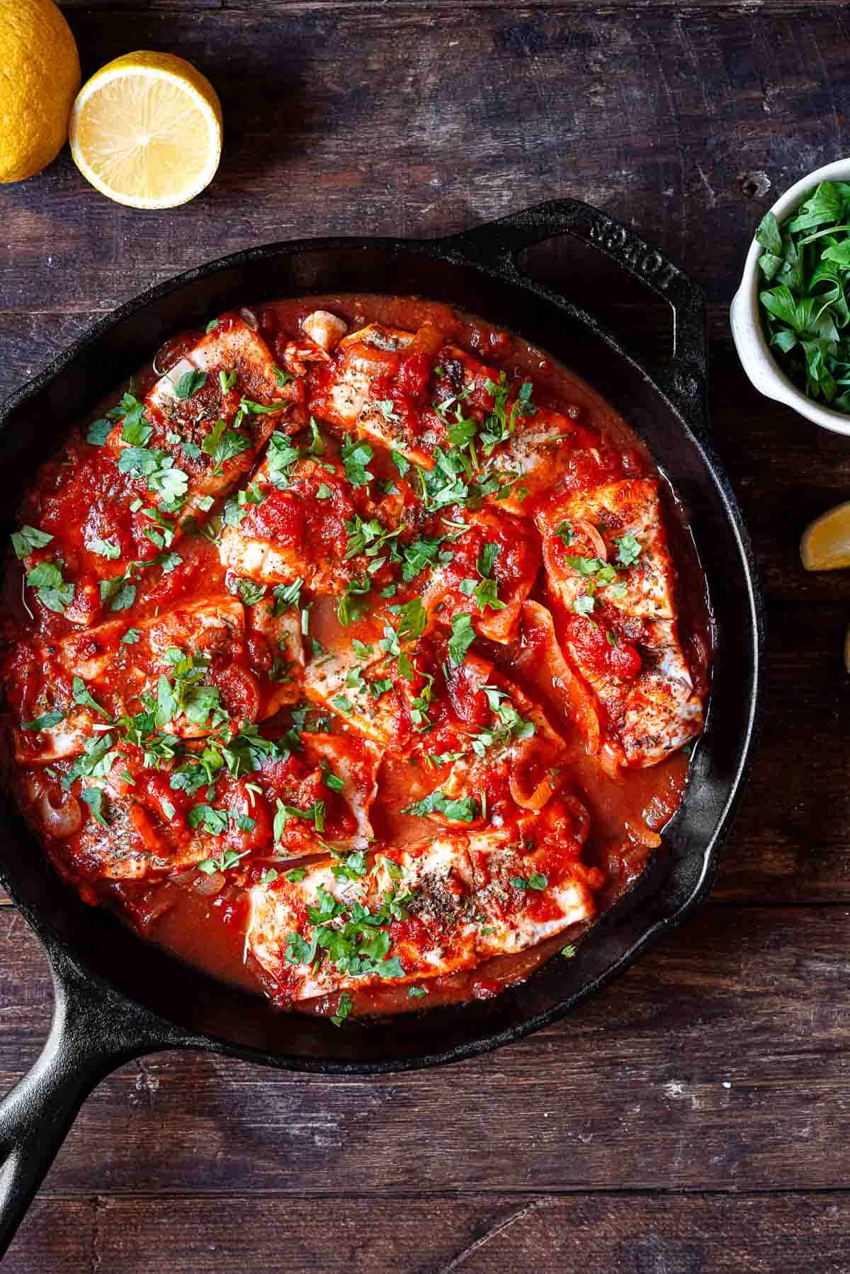 Greek fish with tomatoes and onions in a cast iron pan. parsley and lemon wedges to the side for serving