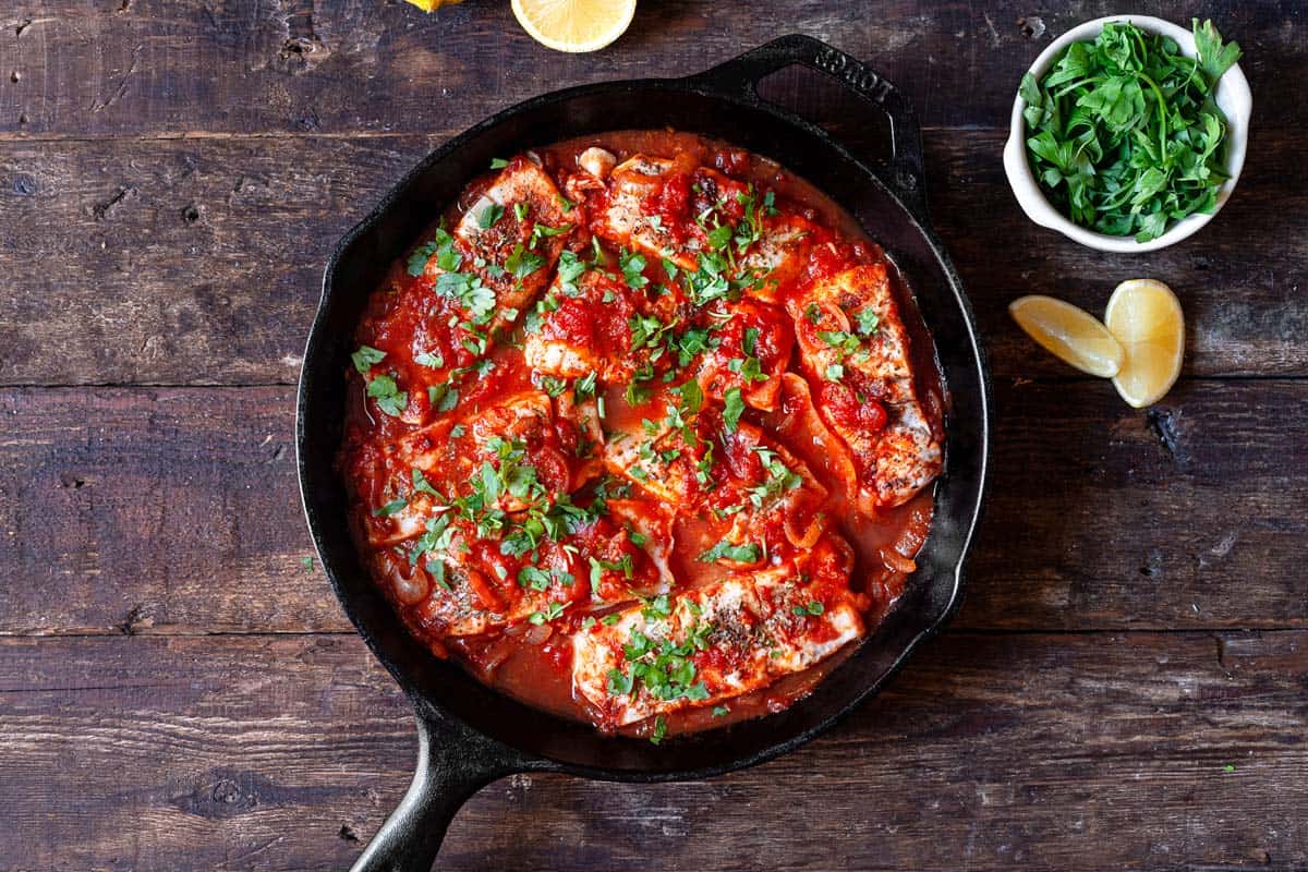 8 pieces of white fish sautéing in a tomato sauce in a cast iron skillet.