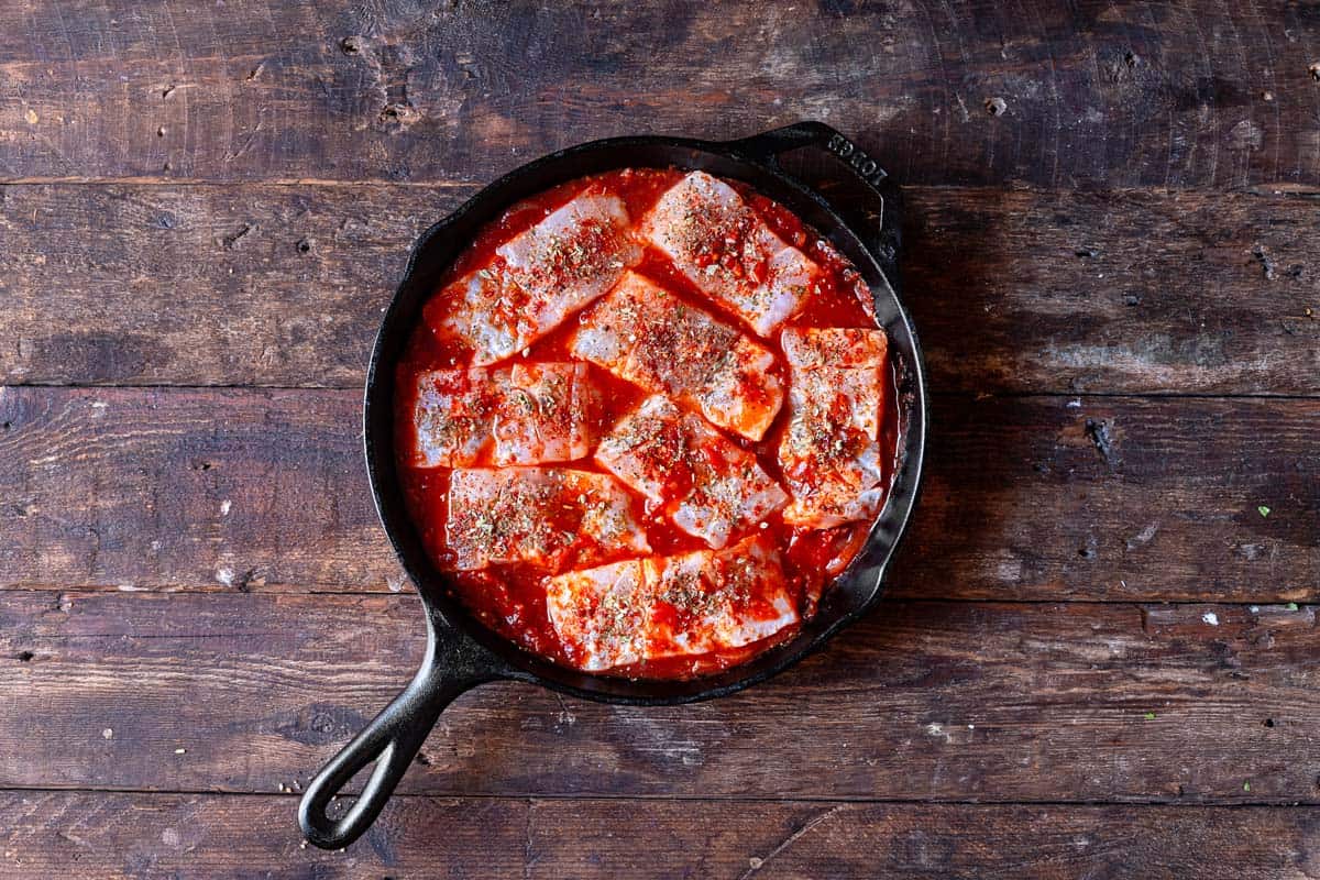 8 pieces of white fish sautéing in a tomato sauce in a cast iron skillet.