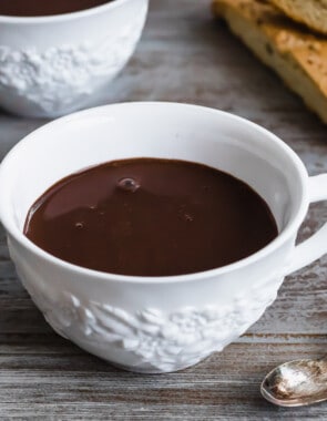 a close up of a mug of hot chocolate next to a spoon and two slices of biscotti.