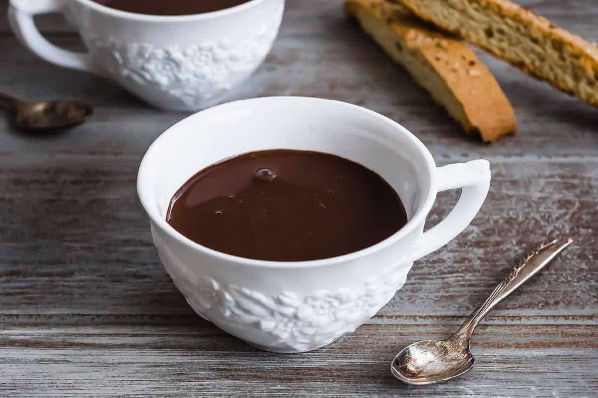 a close up of a mug of hot chocolate next to a spoon and two slices of biscotti.