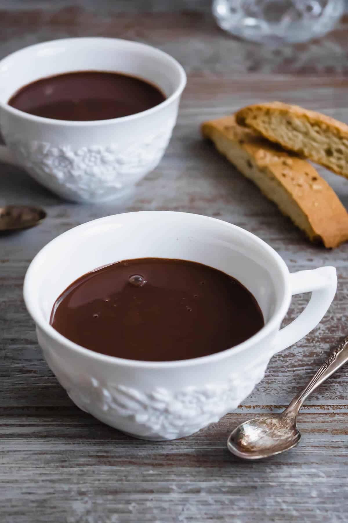 two cups of italian hot chocolate with a spoon and two pieces of biscotti.