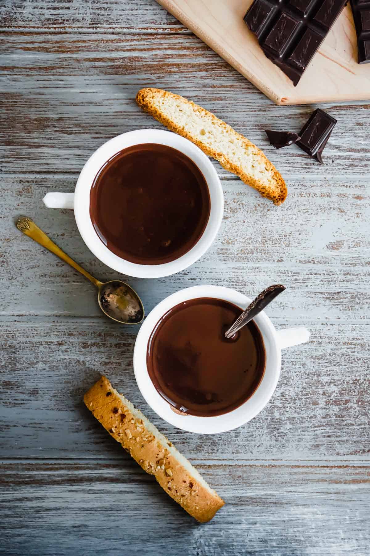 Two cups of Italian hot chocolate (Cioccolato Caldo) with two spoons and two pieces of biscotti.