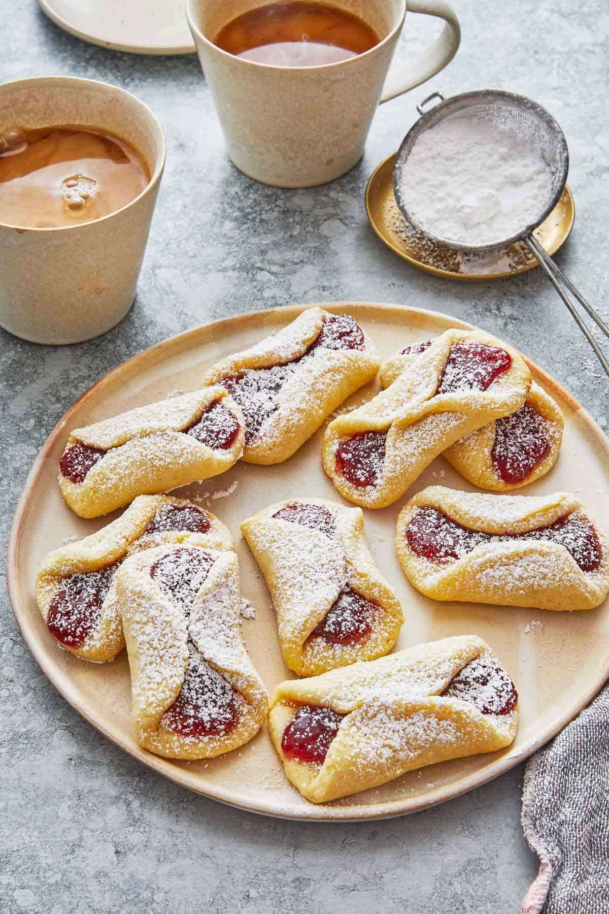 a plate of nine pizzicati cookies with two cups of coffee and a sieve filled with powdered sugar.