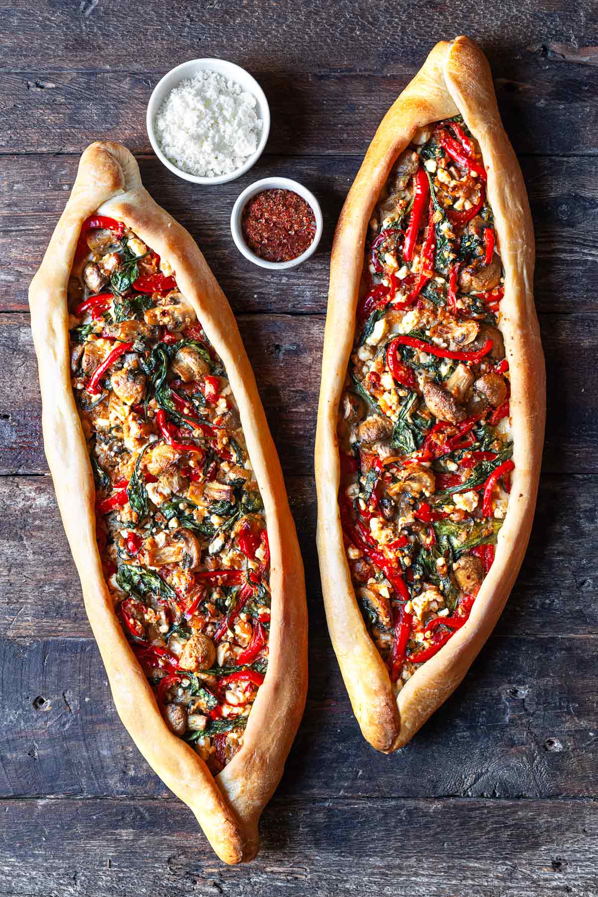 two vegetarian turkish pide on a table with a bowl of aleppo pepper and a bowl of feta cheese.