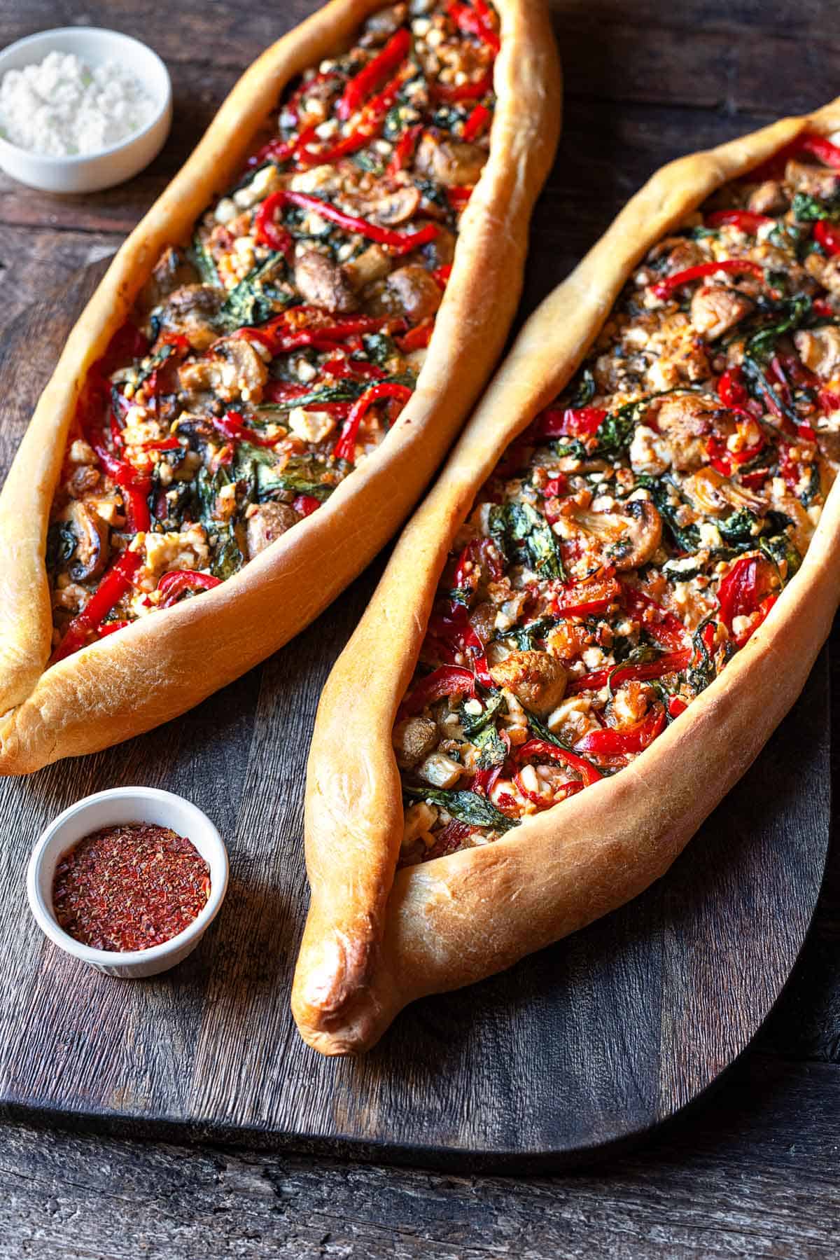 two vegetarian turkish pide on a serving board with a bowl of aleppo pepper and a bowl of feta cheese.