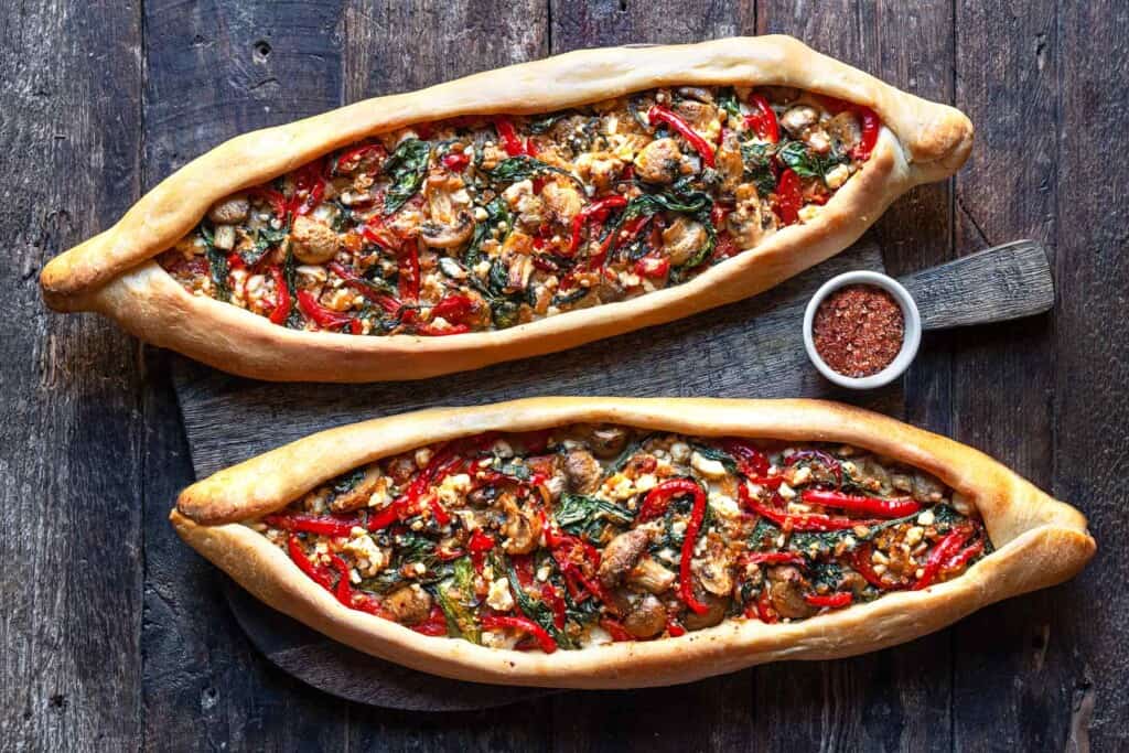 Two baked turkish pide on a serving board with a bowl of aleppo pepper.