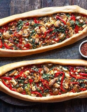 close up of two vegetarian turkish pide on a serving board with a bowl of aleppo pepper.