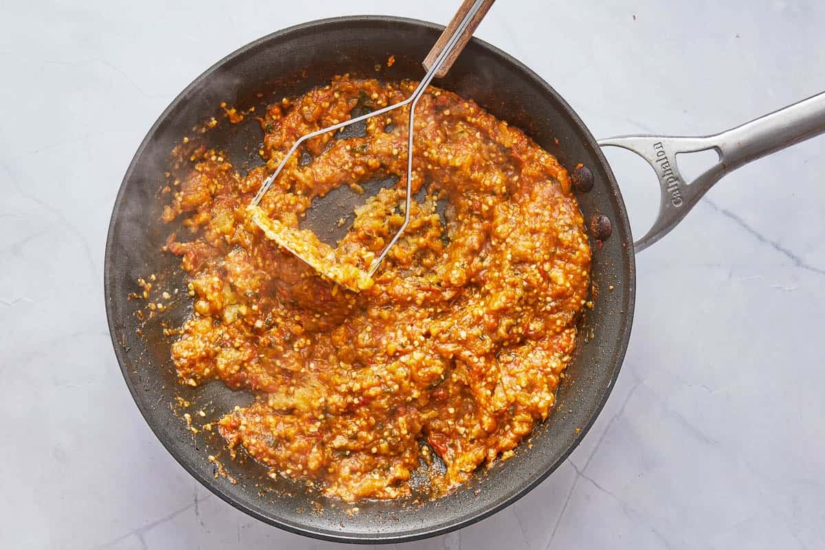 eggplant being mashed with a potato masher in a frying pan.