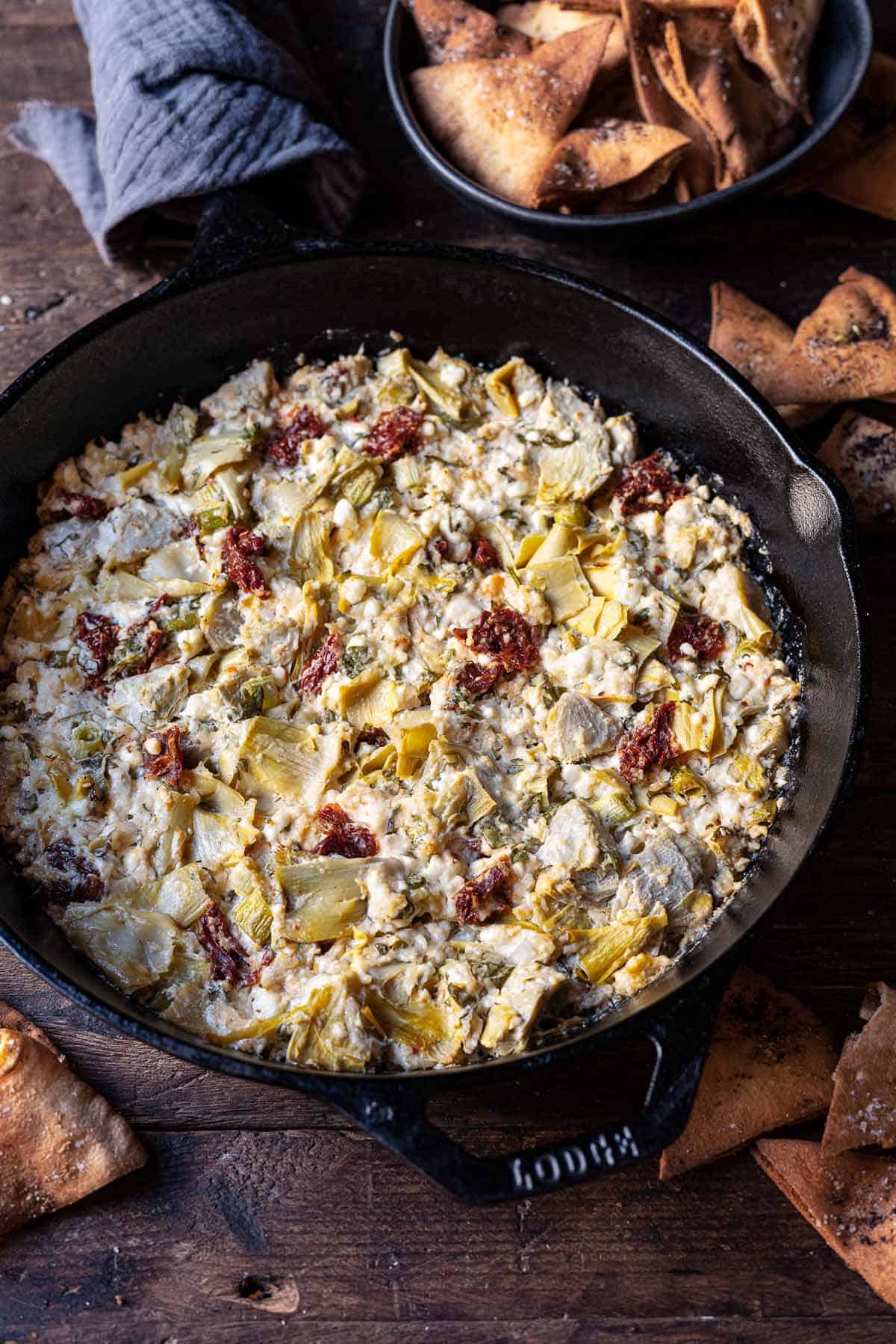 close up of cooked artichoke dip with feta in a cast iron skillet surrounded by fried pita chips.