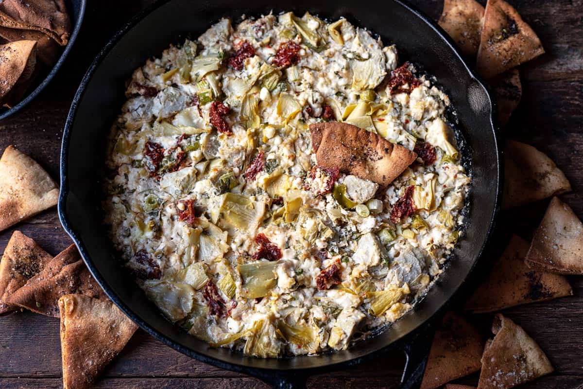 close up of cooked artichoke dip with feta in a cast iron skillet surrounded by fried pita chips.