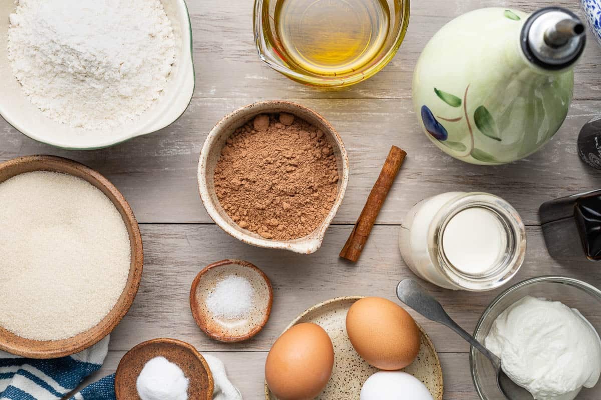 ingredients for chocolate olive oil cake with greek yogurt whip including olive oil, flour, baking soda, salt, cocoa powder, cinnamon, eggs, sugar, heavy cream, and greek yogurt.