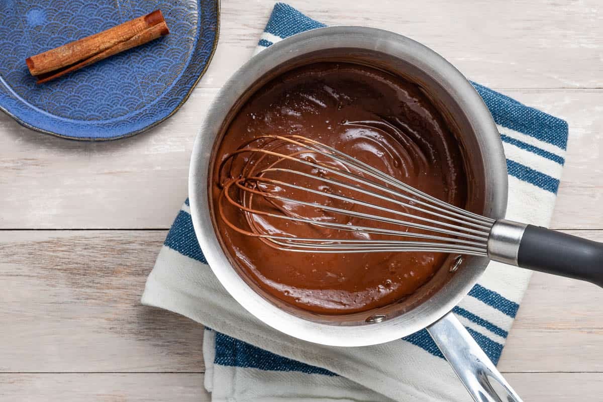 cocoa powder and egg mixtures in a sauce pan with a whisk next to a plate with a cinnamon stick.