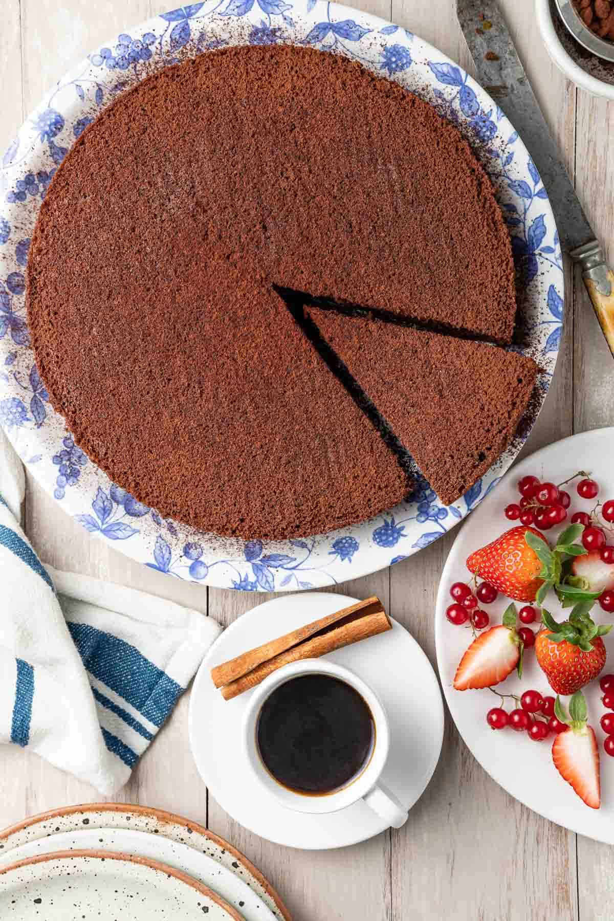 overhead shot of a chocolate olive oil cake on a plate next to a knife, cup of coffee with a cinnamon stick, and a plate of berries.