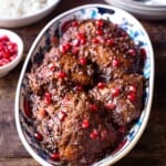 Fesenjan (persian pomegranate and walnut chicken stew) in a serving bowl next to two bowls with spoons, a bowl of rice and a bowl of pomegranate seeds.