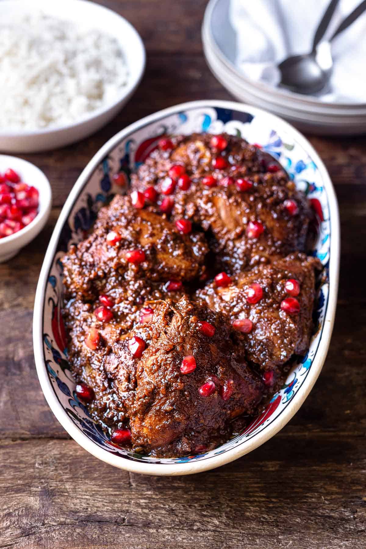 Fesenjan (persian pomegranate and walnut chicken stew) in a serving bowl next to two bowls with spoons, a bowl of rice and a bowl of pomegranate seeds.