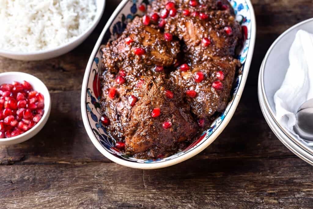 Fesenjan (persian pomegranate and walnut chicken stew) in a serving bowl next to a bowl of rice and a bowl of pomegranate seeds.