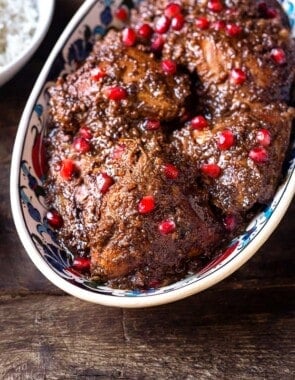 Fesenjan (persian pomegranate and walnut chicken stew) in a serving bowl next to a bowl of rice and a bowl of pomegranate seeds.