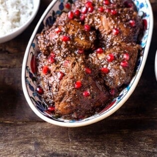 Fesenjan (persian pomegranate and walnut chicken stew) in a serving bowl next to a bowl of rice and a bowl of pomegranate seeds.