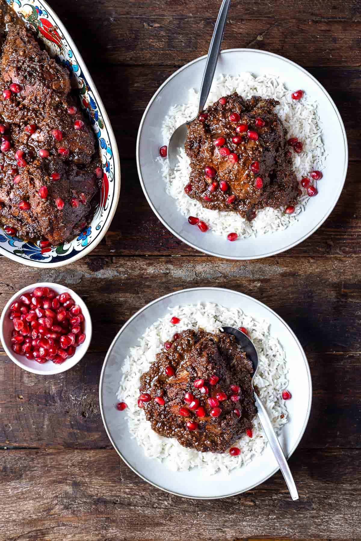 Fesenjan (persian pomegranate and walnut chicken stew) over rice in two bowls with a spoons next to a platter of fesenjan and a bowl of pomegranate seeds.