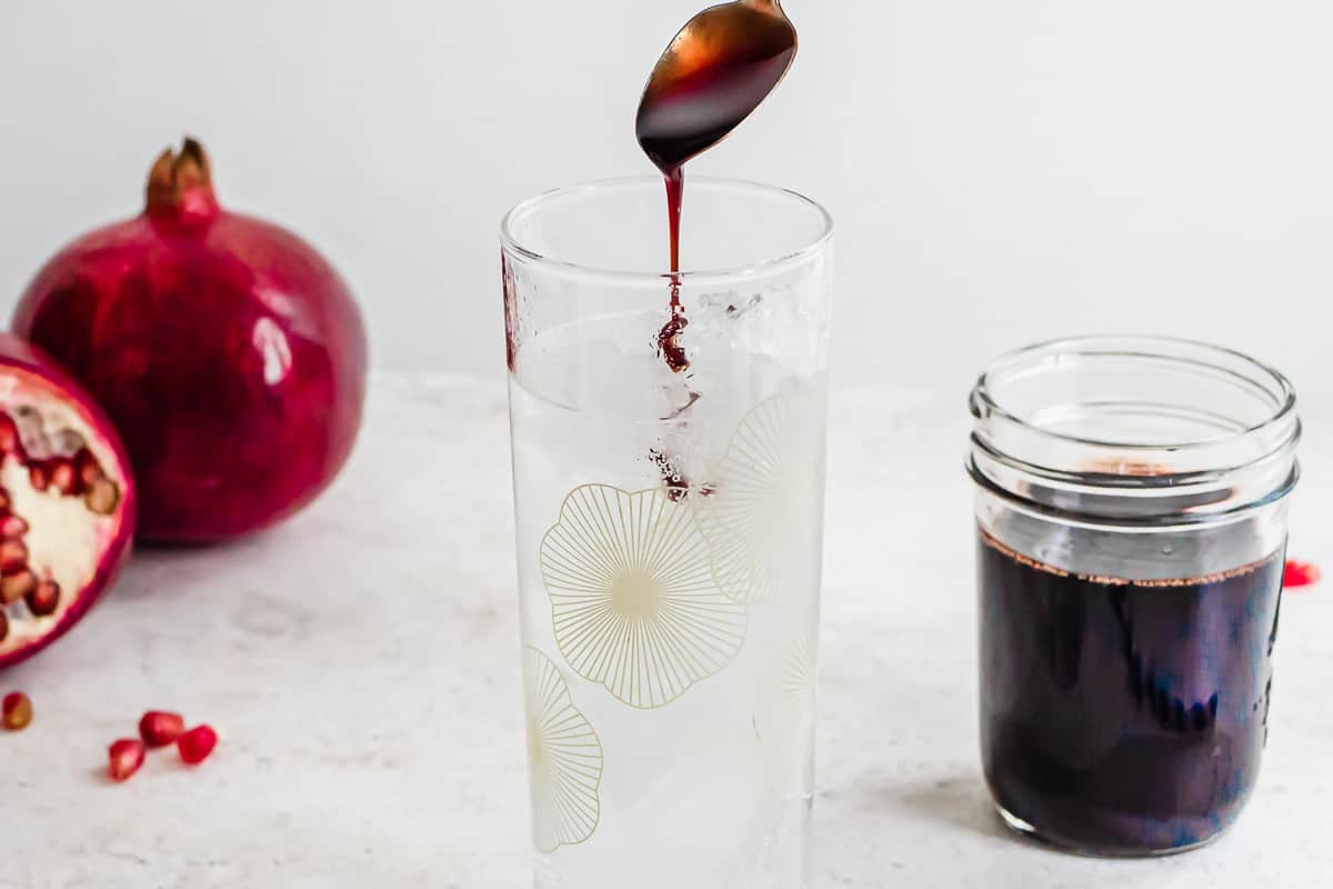 a spoonful of pomegranate molasses being drizzled into a glass of sparkling water next to a jar of pomegranate molasses.