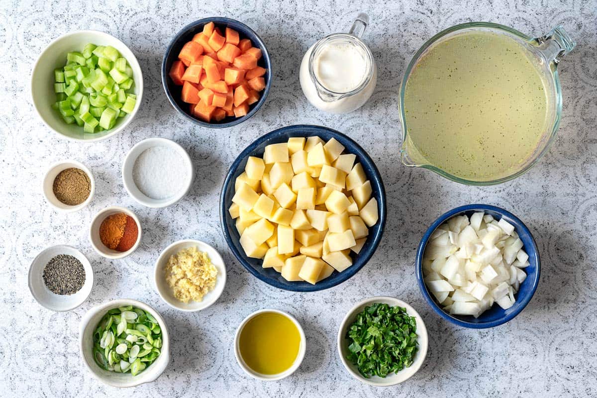 the ingredients for potato soup including olive oil, chopped onion, chopped carrot, chopped celery, minced garlic, diced potatoes, salt, pepper, cumin, paprika, turmeric, vegetable broth, parsley and green onions.