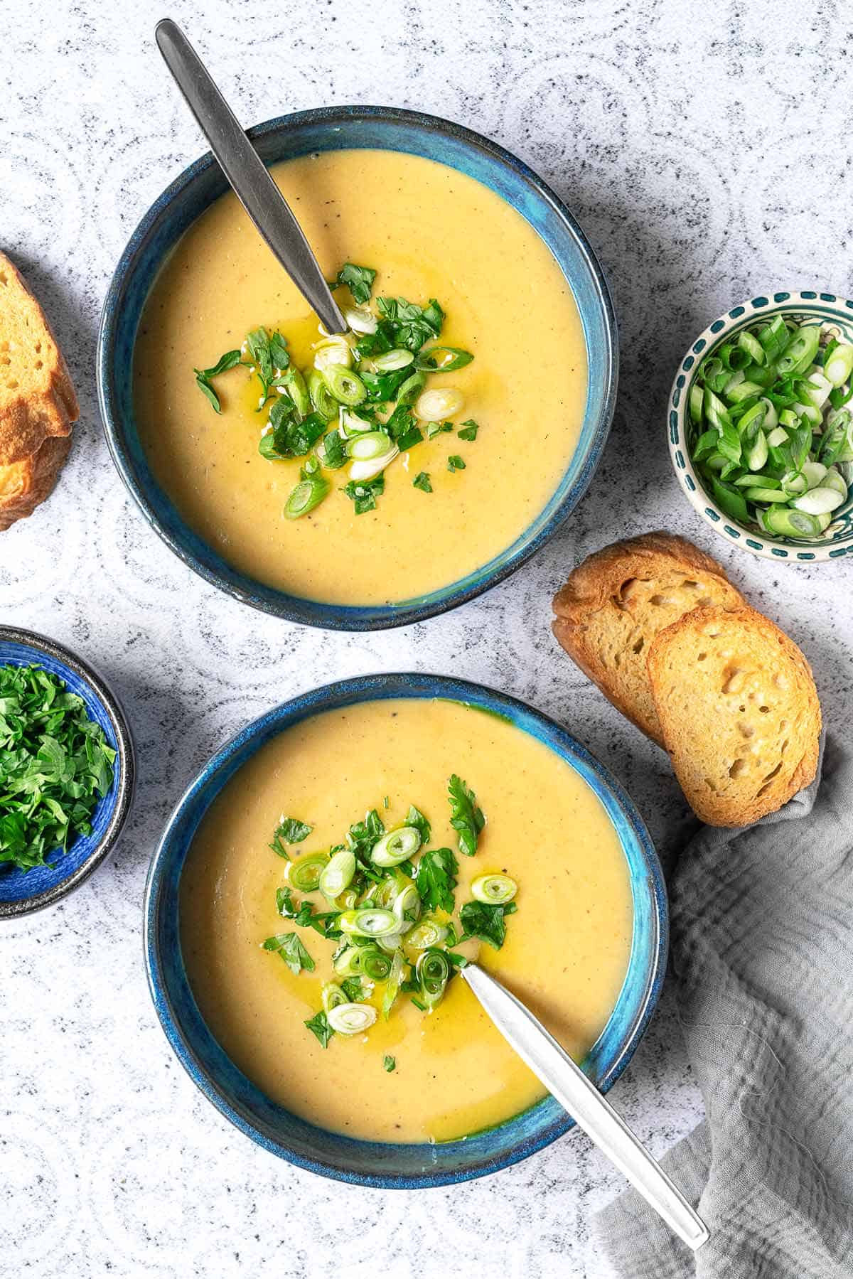 overhead shot of two bowls of potato soup with spoons topped with green onions and parsley next to a bowl of parsley, a bowl of green onions, and two slices of toasted bread.