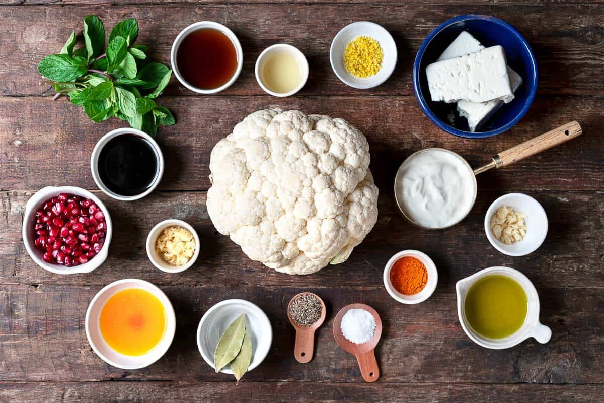 ingredients for whole roasted cauliflower including a head of cauliflower, saffron, kosher salt, turmeric, bay leaves, garlic, olive oil, feta, greek yogurt, lemon zest, lemon juice, pepper, pomegranate molasses, maple syrup, pomegranate seeds and fresh mint leaves.