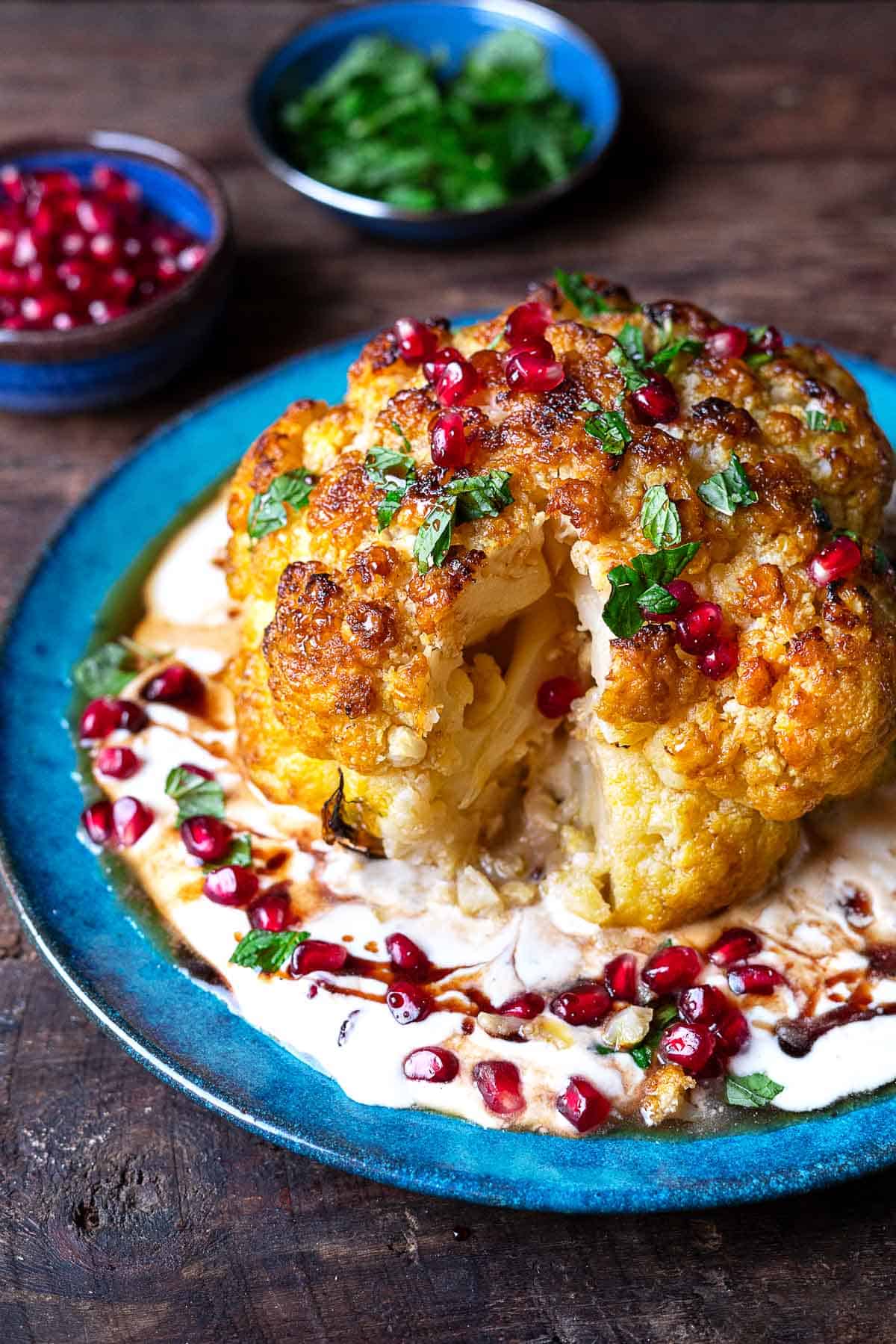 a whole roasted cauliflower with feta and yogurt sauce topped with pomegranate dressing and mint leaves on a plate with a piece cut out of it.