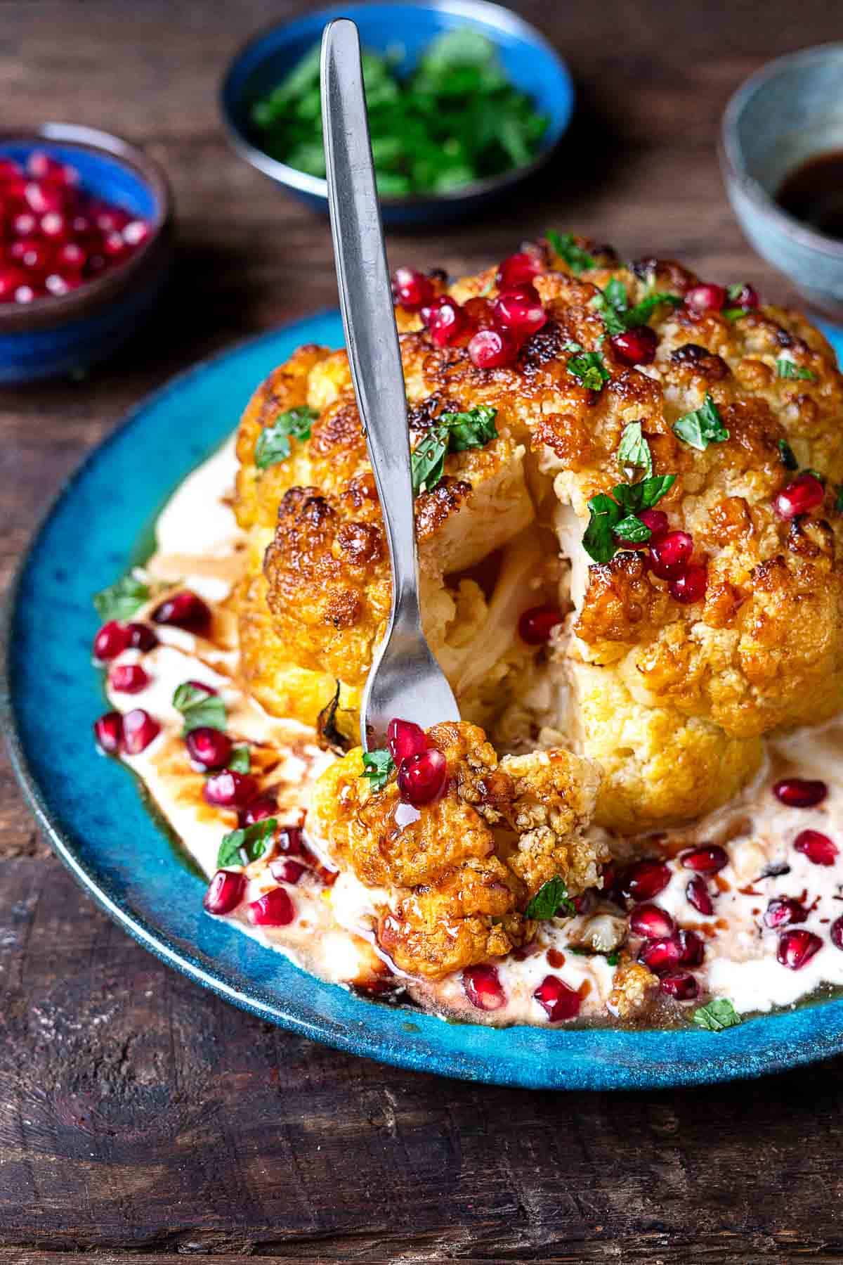 a whole roasted cauliflower with feta and yogurt sauce topped with pomegranate dressing and mint leaves on a plate being cut into by a fork.