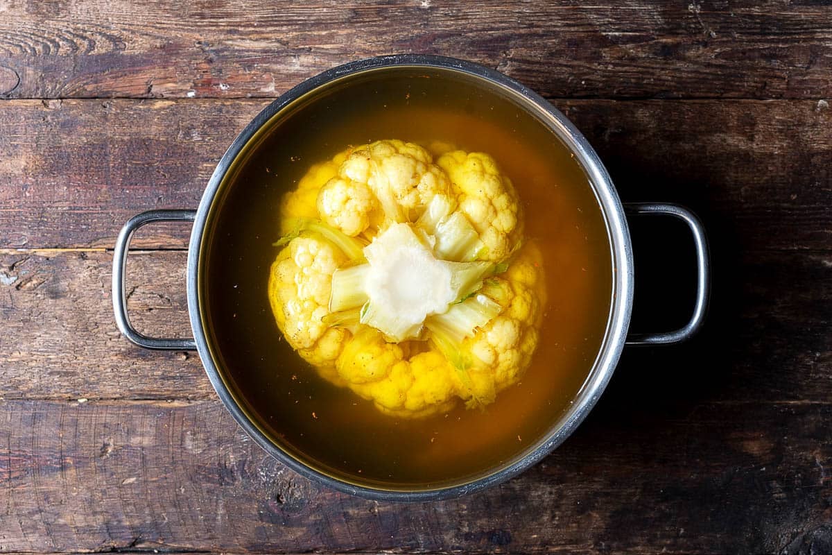 a head of cauliflower boiling in water with kosher salt, turmeric, bloomed saffron, bay leaves and chopped garlic.