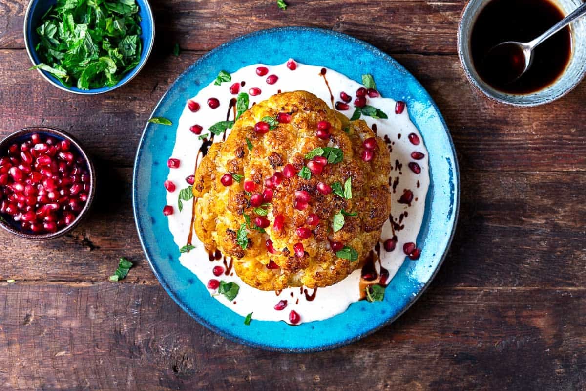 an overhead shot of a whole roasted cauliflower with feta and yogurt sauce topped with pomegranate dressing and mint leaves on a plate.